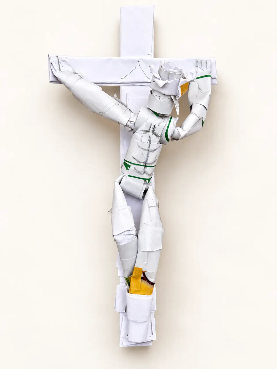 A white paper sculpture by Eklenam of Jesus on the cross, made from folded and rolled paper with small green and yellow details, on a cream background.
