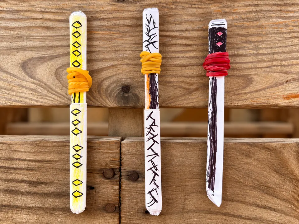 Three paper swords hanging on a wooden fence: one yellow with black diamonds, one white with black patterns and an orange band, and one black and white with a red band.